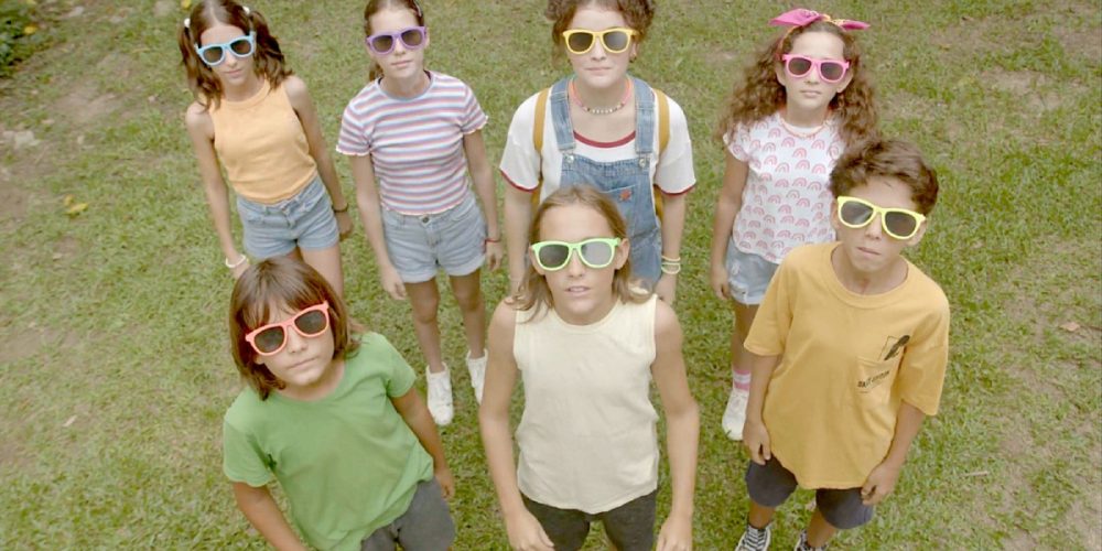 Cena do filme O Cemitério do Parque da Luz mostra seis crianças usam óculos coloridos e olham para cima em um gramado verde, posando em grupo.
