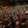 Um auditório lotado de espectadores assistindo a uma apresentação. A plateia, composta por pessoas de diferentes idades, está sentada em cadeiras acolchoadas, em um ambiente iluminado por luzes quentes nas paredes laterais. O foco está no público atento.