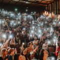 Plateia lotada em teatro escuro, com o público levantando celulares acesos, criando pontos de luz.