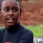 Fotograma de um filme ou série com duas jovens conversando em ambiente externo. Em primeiro plano, uma jovem de cabelos cacheados presos e expressão tensa olha para a outra, parcialmente visível à direita, sugerindo um momento de confronto ou surpresa. Ao fundo, há gramado e terra avermelhada desfocada.