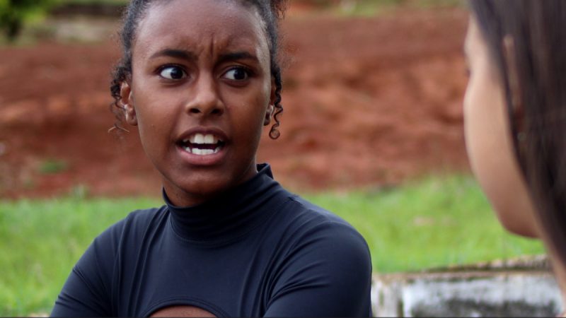 Fotograma de um filme ou série com duas jovens conversando em ambiente externo. Em primeiro plano, uma jovem de cabelos cacheados presos e expressão tensa olha para a outra, parcialmente visível à direita, sugerindo um momento de confronto ou surpresa. Ao fundo, há gramado e terra avermelhada desfocada.
