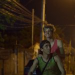 Dois jovens caminham à noite por uma rua iluminada, um de regata verde e boné carregando nas costas outro de regata vermelha que sorri.