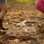 Fotografia de duas pessoas brincando na lama; uma delas salta com botas brancas em uma poça de barro, enquanto a outra caminha com botas pretas, ambas com as pernas sujas.