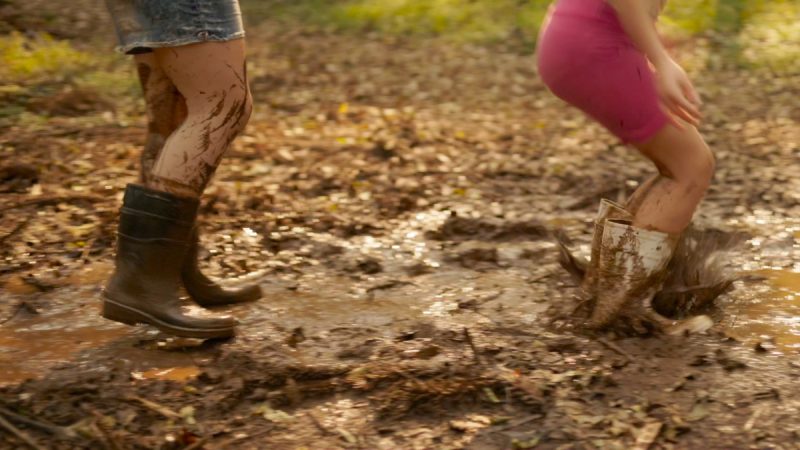 Fotografia de duas pessoas brincando na lama; uma delas salta com botas brancas em uma poça de barro, enquanto a outra caminha com botas pretas, ambas com as pernas sujas.