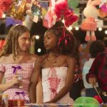 Cena de festa junina com três jovens em frente a uma barraca de jogos; duas usam vestidos caipiras com laços e conversam sob uma fileira de bichos de pelúcia pendurados, enquanto a terceira, de camisa xadrez, participa da pescaria.