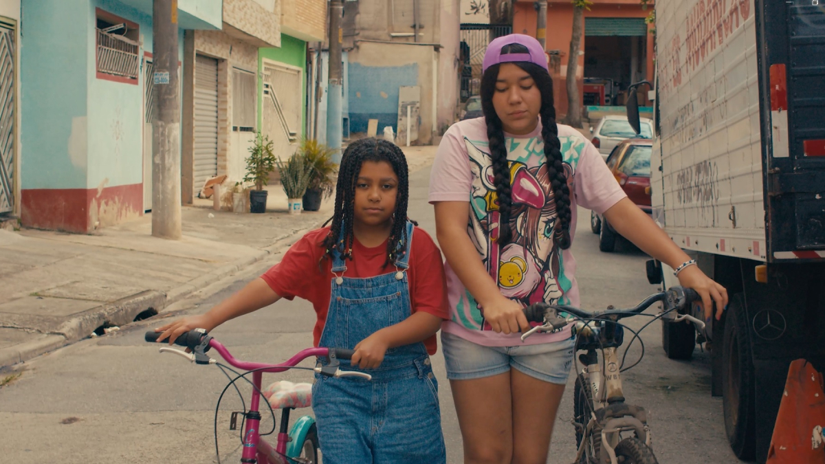 Duas meninas caminham lado a lado empurrando bicicletas por uma rua de bairro residencial; a menor usa camiseta vermelha e jardineira jeans, enquanto a outra veste camiseta estampada, boné roxo e shorts, ambas com expressão séria.