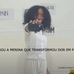 Cena de vídeo com menina de cabelos cacheados e roupa branca segurando um frasco de tinta preta à frente; ao fundo, palavras como “PAIN” e “STRENGTH” coladas na parede e a frase central em destaque: “SOU A MENINA QUE TRANSFORMOU DOR EM PODER”.