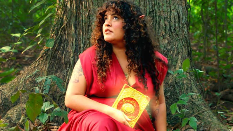 Mulher com orelhas de elfa e vestido vermelho sentada ao pé de uma árvore em floresta, segurando um livro dourado brilhante no colo e olhando para cima com expressão contemplativa.