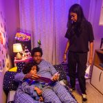 Homem e criança de pijama sentados na cama leem um livro, enquanto adolescente em pé observa no quarto decorado com temática infantil.