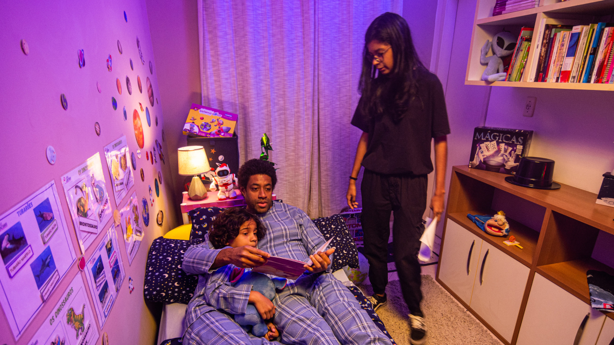 Homem e criança de pijama sentados na cama leem um livro, enquanto adolescente em pé observa no quarto decorado com temática infantil.