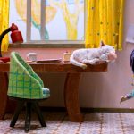 Cenário de animação em stop motion com uma criança de papel machê ao lado de uma mesa, onde um gato branco dorme; o quarto é colorido, com cortinas amarelas, desenhos infantis na parede e objetos escolares sobre a mesa.