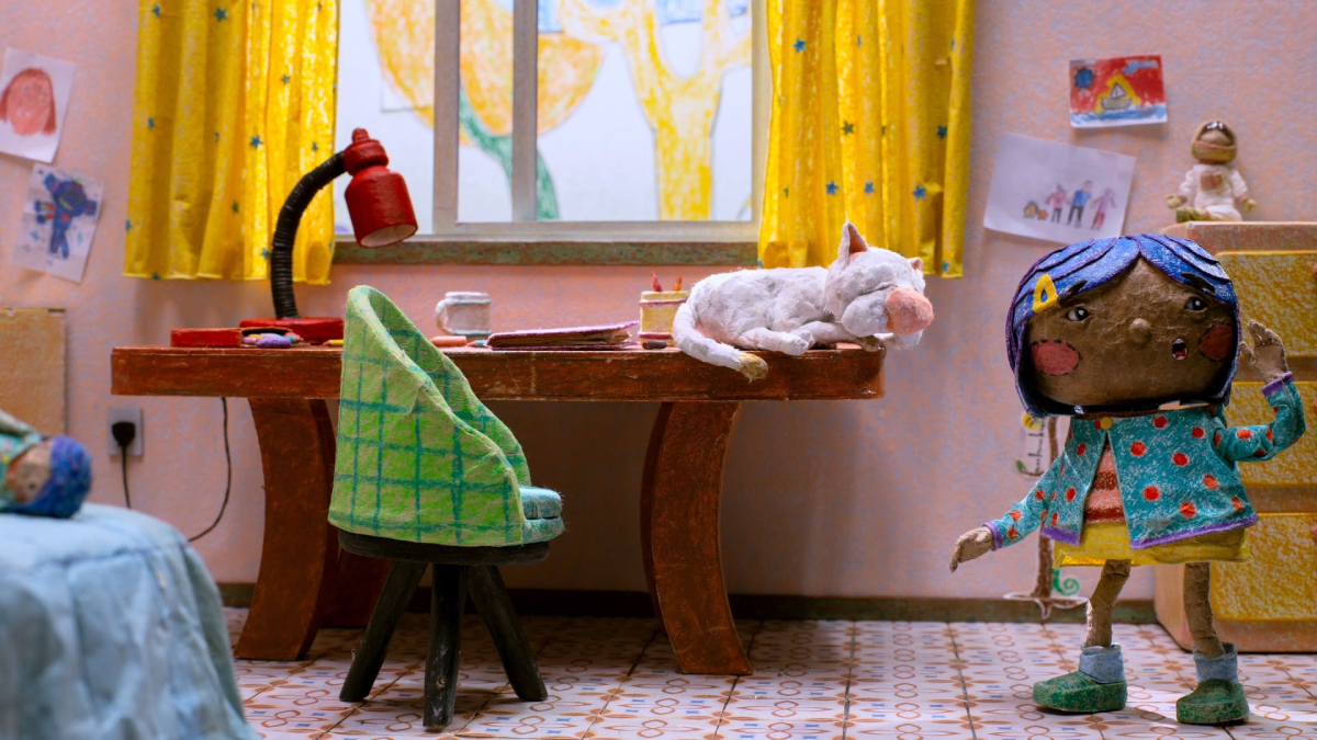 Cenário de animação em stop motion com uma criança de papel machê ao lado de uma mesa, onde um gato branco dorme; o quarto é colorido, com cortinas amarelas, desenhos infantis na parede e objetos escolares sobre a mesa.