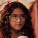 Menina de cabelos cacheados e óculos grandes olha atentamente, com laço rosa no cabelo, em ambiente interno iluminado suavemente.