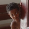 Menino de pele negra e cabelo crespo curto observa com expressão curiosa enquanto espreita por trás de uma porta de madeira entalhada.
