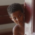 Menino de pele negra e cabelo crespo curto observa com expressão curiosa enquanto espreita por trás de uma porta de madeira entalhada.