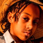 Criança com tranças usa chapéu de palha e camisa listrada, com bigode desenhado no rosto, olhando diretamente para a câmera.