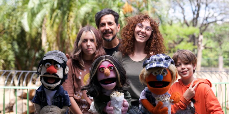 Grupo sorridente com dois adultos, duas crianças e três fantoches coloridos segurando saquinhos de pipoca, em ambiente externo arborizado.