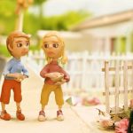 Animação em massinha mostra dois bonecos crianças caminhando na calçada com cestas de pães, enquanto um idoso em cadeira de rodas aparece à direita.