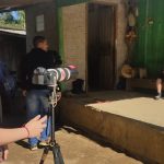 Jovem com blusa vermelha opera uma câmera profissional em tripé, enquanto um menino sentado em banco de madeira é filmado em frente a uma casa simples com parede verde.