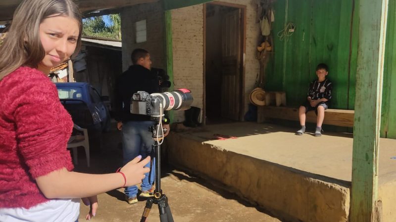 Jovem com blusa vermelha opera uma câmera profissional em tripé, enquanto um menino sentado em banco de madeira é filmado em frente a uma casa simples com parede verde.