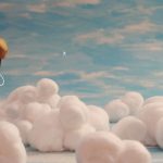 Figura estilizada com cabeça arredondada marrom e amarela e corpo simples desenhado flutua sobre nuvens feitas de algodão, com céu azul ao fundo.