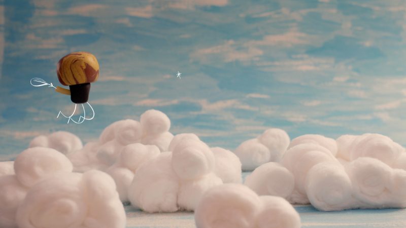 Figura estilizada com cabeça arredondada marrom e amarela e corpo simples desenhado flutua sobre nuvens feitas de algodão, com céu azul ao fundo.