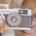 Pessoa segura uma câmera analógica Olympus Trip 35 próxima ao rosto, sorrindo com um dos olhos fechados enquanto enquadra a foto.