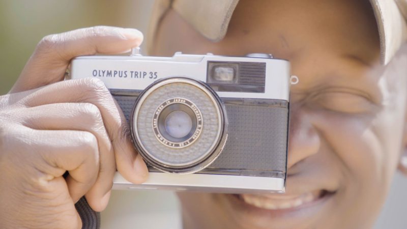 Pessoa segura uma câmera analógica Olympus Trip 35 próxima ao rosto, sorrindo com um dos olhos fechados enquanto enquadra a foto.