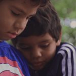 Dois meninos estão próximos, um vestindo camiseta azul com estampa circular vermelha e branca, e o outro usando agasalho azul-marinho com listras brancas, ambos com expressão concentrada.