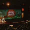 Fotografia de um auditório durante a 23ª Mostra de Cinema Infantil de Florianópolis, com quatro pessoas no palco diante de uma grande tela colorida que exibe a identidade visual do evento.