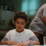 Menino de camiseta branca com listras nas mangas está concentrado em atividade sobre a mesa, enquanto um homem mais velho, ao fundo, cozinha em uma cozinha simples.