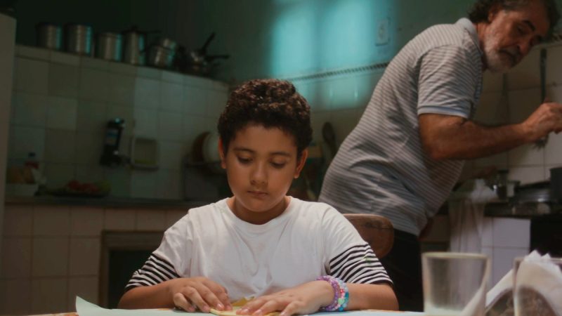 Menino de camiseta branca com listras nas mangas está concentrado em atividade sobre a mesa, enquanto um homem mais velho, ao fundo, cozinha em uma cozinha simples.