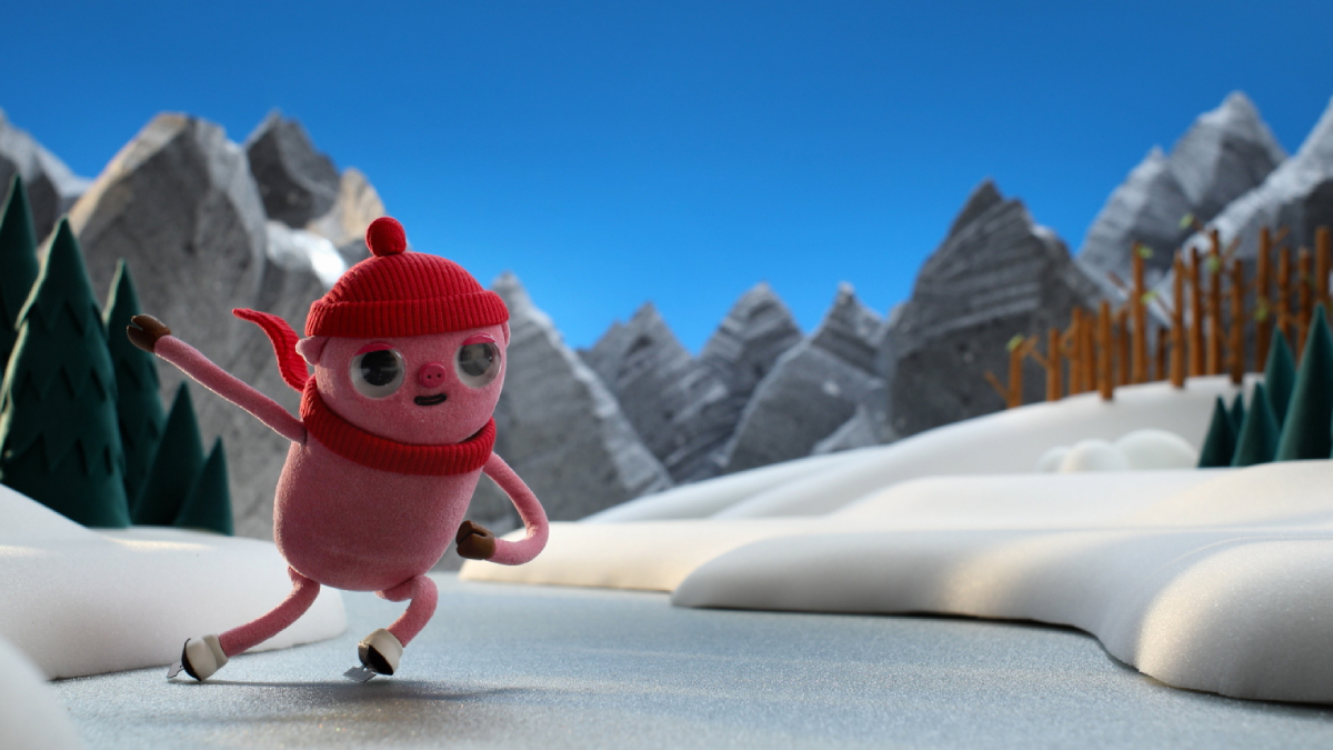 Animação em stop motion mostra um personagem rosado com olhos grandes, gorro e cachecol vermelhos patinando no gelo em meio a uma paisagem de neve, pinheiros verdes e montanhas cinzentas ao fundo sob céu azul.