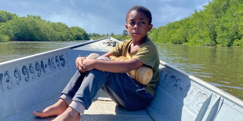 Menino sentado descalço na proa de um barco metálico, com expressão séria, cercado por águas calmas e vegetação verde.
