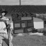 Fotografia em preto e branco mostra duas crianças encenando um faroeste. À esquerda, um menino veste chapéu de cowboy, lenço no ombro e calça estampada, em frente a uma construção simples. À direita, outro menino segura uma claquete artesanal que traz escrito “El bueno, el malo y el feo”, “Director: Sergio Leone”, além de indicações de tomada, cena e som.