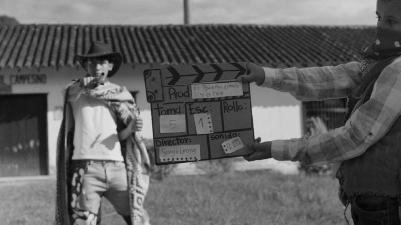 Fotografia em preto e branco mostra duas crianças encenando um faroeste. À esquerda, um menino veste chapéu de cowboy, lenço no ombro e calça estampada, em frente a uma construção simples. À direita, outro menino segura uma claquete artesanal que traz escrito “El bueno, el malo y el feo”, “Director: Sergio Leone”, além de indicações de tomada, cena e som.