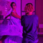 Mulher de vestido esvoaçante dança sorridente sob luz roxa intensa, enquanto outra pessoa a observa em ambiente com mapas na parede e cadeiras escolares.