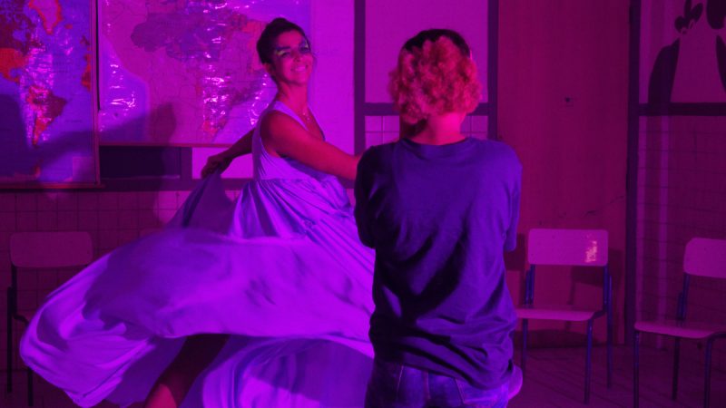 Mulher de vestido esvoaçante dança sorridente sob luz roxa intensa, enquanto outra pessoa a observa em ambiente com mapas na parede e cadeiras escolares.