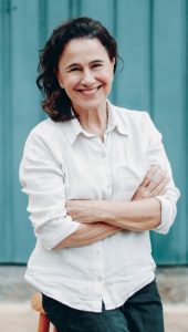 Mulher de cabelos castanhos ondulados, vestindo camisa branca de mangas dobradas, sorri com os braços cruzados diante de um fundo azul.