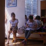 Fotografia mostra duas meninas sentadas em um banco de madeira dentro de uma sala iluminada pela luz natural que entra pela janela gradeada. À esquerda, uma menina veste collant branco e sapatilhas de balé, olhando para frente. À direita, outra menina com camiseta estampada e laço colorido no cabelo escreve em um caderno apoiado no colo. Ao fundo, outras crianças aparecem parcialmente.