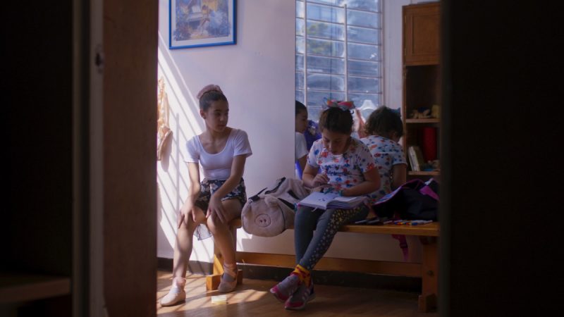 Fotografia mostra duas meninas sentadas em um banco de madeira dentro de uma sala iluminada pela luz natural que entra pela janela gradeada. À esquerda, uma menina veste collant branco e sapatilhas de balé, olhando para frente. À direita, outra menina com camiseta estampada e laço colorido no cabelo escreve em um caderno apoiado no colo. Ao fundo, outras crianças aparecem parcialmente.