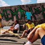 Cinco jovens posam em rua diante de muro com grafite “Memória de Pivete”, três vestindo camisas da seleção brasileira e um segurando pipa com estampa da bandeira.