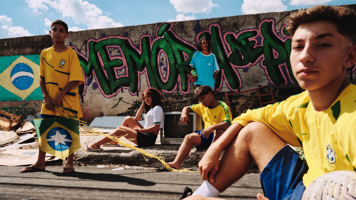 Cinco jovens posam em rua diante de muro com grafite “Memória de Pivete”, três vestindo camisas da seleção brasileira e um segurando pipa com estampa da bandeira.