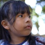 Menina de cabelos pretos e longos com franja segura as correntes de um balanço, olhando para o lado com expressão atenta.