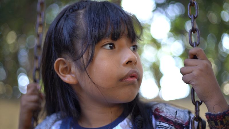 Menina de cabelos pretos e longos com franja segura as correntes de um balanço, olhando para o lado com expressão atenta.