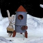 Cena em stop motion mostra um coelho antropomórfico de pelúcia, com bolsa a tiracolo, ao lado de um pequeno foguete metálico em uma paisagem lunar de gelo e rochas, com o céu estrelado ao fundo.