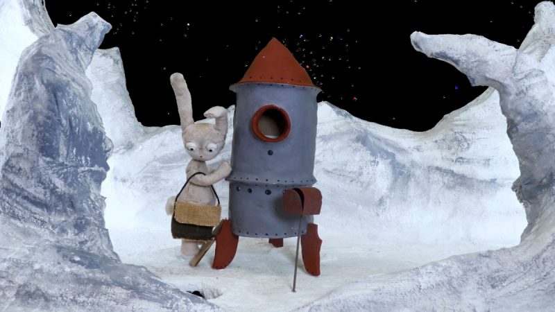 Cena em stop motion mostra um coelho antropomórfico de pelúcia, com bolsa a tiracolo, ao lado de um pequeno foguete metálico em uma paisagem lunar de gelo e rochas, com o céu estrelado ao fundo.