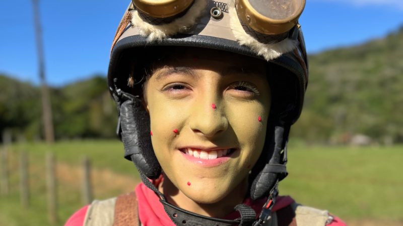 Menino sorridente com o rosto pintado de verde e pequenas bolinhas vermelhas, usando capacete com óculos de aviador presos e mochila nas costas, em ambiente de campo ensolarado.