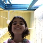 Menina sorridente de cabelos castanhos curtos olha para a câmera de baixo para cima, em ambiente interno iluminado por claraboia azul no teto e paredes claras ao redor.