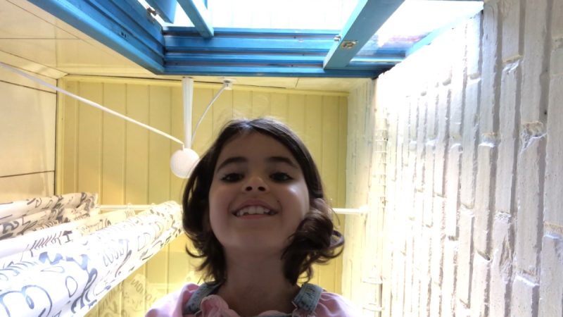 Menina sorridente de cabelos castanhos curtos olha para a câmera de baixo para cima, em ambiente interno iluminado por claraboia azul no teto e paredes claras ao redor.